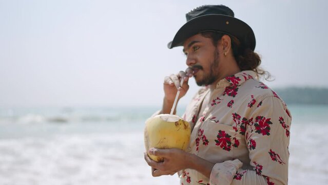 Vibrant LGBTQ man cowboy sips coconut on sunny beach. Flamboyant style, ocean backdrop, holiday vibe. Confident individual enjoys tropical drink, waves roll in. Pride, relaxation meet seaside leisure.