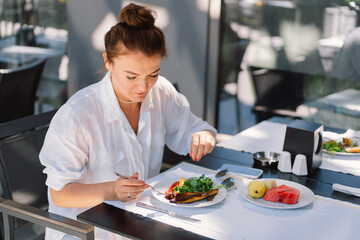 A woman in a white shirt eats lunch or breakfast outdoors in a cafe. Woman eating healthy food on a restaurant terrace