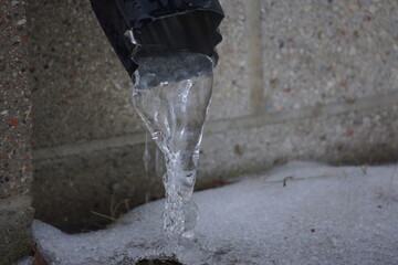 Macro of black water spout with frozen water flowing out, grey brick background, snow on ground