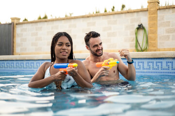 A couple of different ethnicities are playing in the pool with water guns. The young people are...