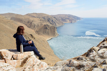 A young female traveler is sitting on a rock overlooking a spring lake and drinking hot tea from a...