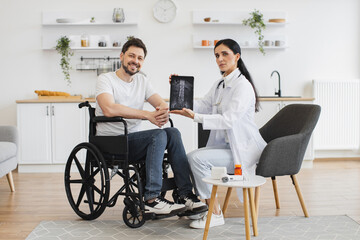 Portrait of young female nurse and mature man wheelchair user, showing x-ray scan of spine on modern tablet. Caucasian woman doctor checking up condition of mature bearded male patient at home.