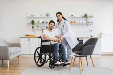 Full length portrait of female doctor hugging smiling male patient with disability posing at home at background of light kitchen. Health professional giving recommendation and care about treatment .