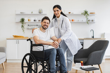 Portrait of female doctor hugging smiling male patient with disability posing at home at background of light kitchen. Health professional giving recommendation and care about treatment .