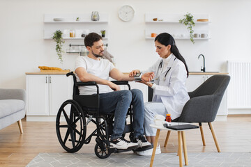 Obraz premium Adult man in wheelchair listening explanations about necessity of leading healthy lifestyle. Attractive cardiologist telling patient with disability about all reasons of hypertension sitting at home.