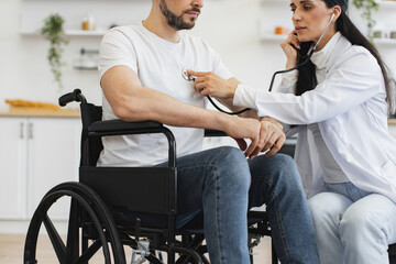 Naklejka premium Cropped view of focused lady in white coat using stethoscope while mature man in casual clothes sitting on wheelchair. General practitioner examining heartbeat while providing regular checkup at home.