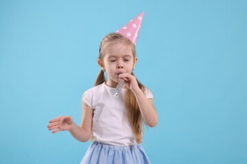 Birthday celebration. Cute little girl in party hat with blower on light blue background