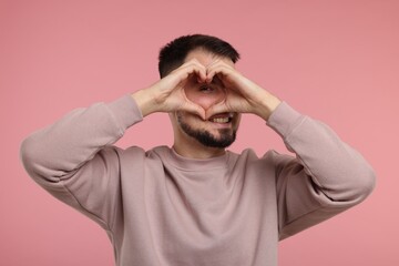 Man looking through folded in shape of heart hands on pink background