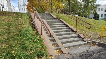 Concrete tile stairs with metal railings lead from the city park to the city block. Nearby are grassy lawns with fallen leaves, trees with yellowed leaves, and buildings. Autumn sunny weather