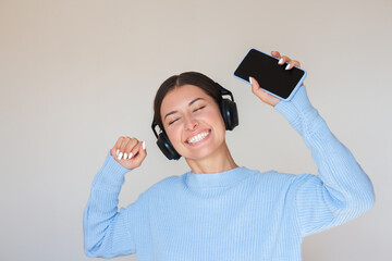 A young pretty brunette woman in a blue sweater listens to music in large black wireless headphones, holds a phone in her hand, closes her eyes, smiles and dances. Isolated on a beige background