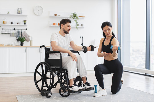 Physiotherapist in sportswear showing adult man in wheelchair how do exercises with dumbbells at home. Male patient undergoing rehabilitation after illness or injury in background of modern apartment. - Powered by Adobe