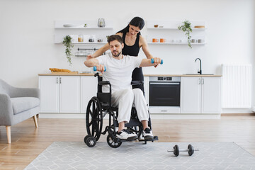 Nurse or wife helping mature man in wheelchair lifting arms with dumbbells for muscle strength in rehabilitation. Physiotherapy healthcare, medical doctor consulting disabled caucasian patient.