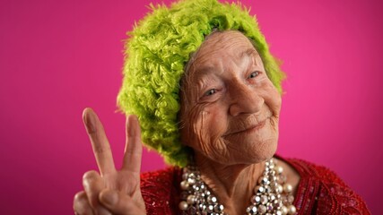 Funny, elderly mature woman, 80s, giving peace sign gesture with no teeth and green hat, isolated...