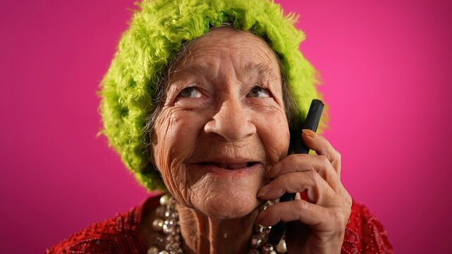 Funny closeup view of toothless old elderly woman wearing green wig or hat, isolated on pink background talking on cell phone.