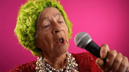 Slow motion of funny closeup fisheye view of elderly woman singing enthusiastically into a microphone and dancing wearing green hat or wig isolated on pink background
