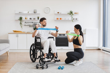 Female physiotherapist or wife showing mature male patient in wheelchair how to exercise with...
