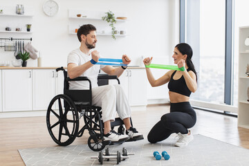 Female physiotherapist or wife showing mature male patient in wheelchair how to exercise with...