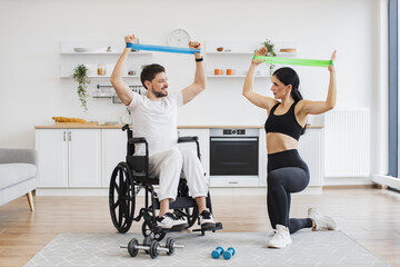 Fototapeta premium Wife in sportswear helping mature man in wheelchair lifting arms with stretching bands for muscle strength. Physiotherapy healthcare, medical trainer consulting disabled caucasian patient.