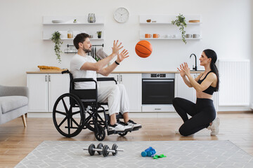 Female physiotherapist or wife throws basketball to young man in wheelchair doing workout exercises at home. Senior male patient undergoing rehabilitation after illness or injury.