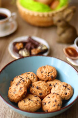 Easter eggs in the basket, bowl of cookies, chocolate pralines, Easter bunny figurine, cups of tea, glasses of juice, flowers and lit candles on the table. Selective focus.