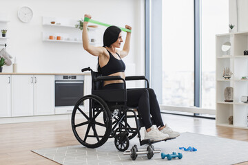 Full length view of Caucasian woman in wheelchair stretching bands while exercising with limited mobility indoors. Smiling adult using moderate-intensity activity during home workout in kitchen.
