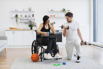 Rehab fitness for senior patients concept. Male physiotherapist or husband showing mature female patient in wheelchair how to exercise with dumbbells at home. - obrazy, fototapety, plakaty