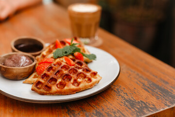Waffles with strawberries and honey on a white plate. Delicious pastries with berries and chocolate for breakfast. Belgian waffles and coffee on table side view