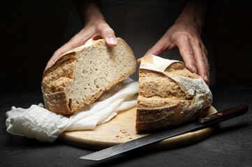 Hands with loaf of bread cut in half on worktop with cutting board, knife and tea towel, close-up.