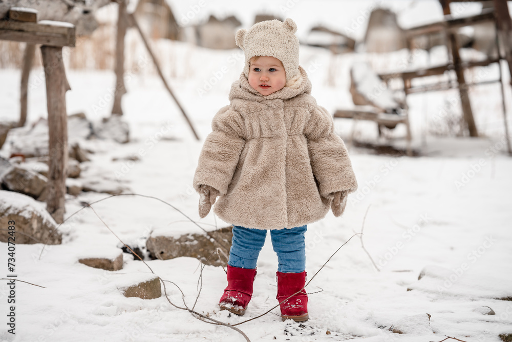 Wall mural cute little baby child toddler in a plush hat and fur coat on a snowy winter landscape with reeds. a