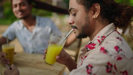 Confident man enjoys orange beverage at tropical cafe. Male with floral shirt sips juice straw, friend in background. Relaxed individuals, casual meet, sunny ambiance. - Powered by Adobe