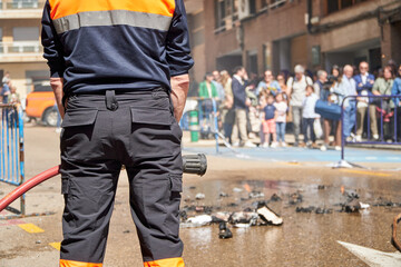 Fireman in with hose staying ready in front of a controlled fire on the street at day time while people look to the wet and smoky floor from the background