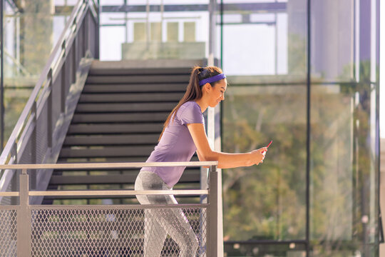 Latino woman in sportswear leaning on the fence of the stairs texting on his cell phone after his workout. Healthy lifestyle. 