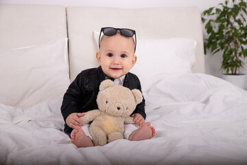 Portrait of smiling child in glasses on head hands holding teddy bear sitting on bed in white room with green flower stylish little kid