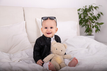 Smiling child european appearance 1 year old with glasses on head watching camera in stylish clothes in white room on bed with green flower