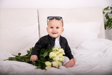 Stylish child  portrait of a boy of european appearance with glasses on his head in a leather jacket holding a rose white in his hands sitting on a white bed Congratulations on March 8, birthday