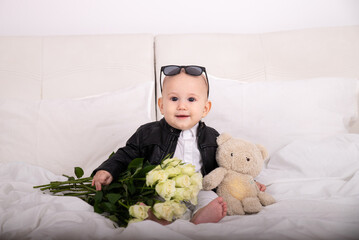 Portrait of a newborn boy 1 year old sitting in stylish clothes with glasses on his head with roses and a teddy bear in his hands White roses baby greeting card March 8 birthday