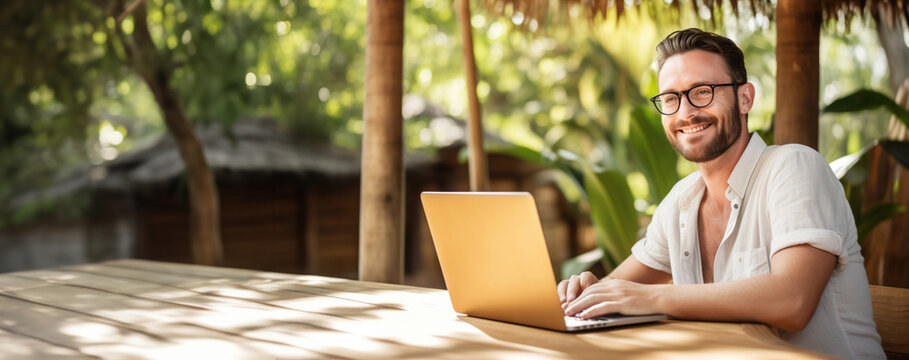 A happy man works remotely on his laptop at a wooden desk, surrounded by lush tropical foliage, in a serene, sunny, natural environment. Digital nomad concept.