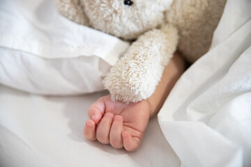 Hand of a little child and a teddy bear toy lying on a white bed on a bed, love for toys, child sleeping