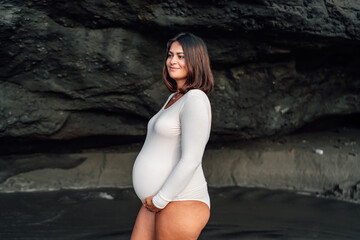 Radiant young pregnant woman, early 30s, smiling gently in a long-sleeve white bodysuit, standing on a sandy beach with a rocky backdrop, capturing a serene maternity moment.