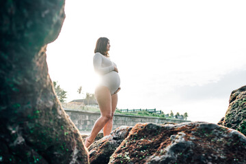 radiant pregnant woman in tight white one-piece stands between rocks on beach, basking in glow of...