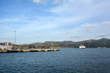 A pier in a sea bay with a ferry sailing away from it under a blue sky