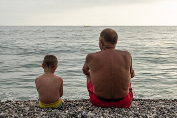 Grandfather and his grandson admiring the sea sitting on the shore. back view