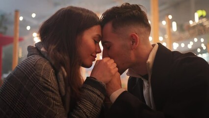 Side view of romantic date of young affectionate couple holding hands sitting at cafe table face to face. Happiness, joy moment, love feelings of two lovers enjoying Christmas evening in city.