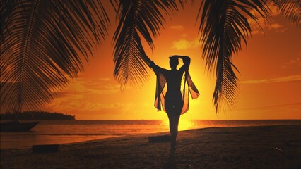 Woman silhouette against orange sunset sky. Young slim woman walking tropical island beach under the coconut palm tree branch. Relaxing, travel, tourism, exotic holiday vacation, back view