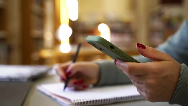 Close-up female hands using smartphone, typing online messaging in sunny room. Professor making notes in notebook, writing thesis, information, create plan or time schedule. Business education study