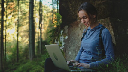 Woman enjoy working in wild nature landscape. Freelance girl typing on laptop while resting sitting under the tree in beautiful forest. Digital nomad remote work IT specialist, people travel, tourism