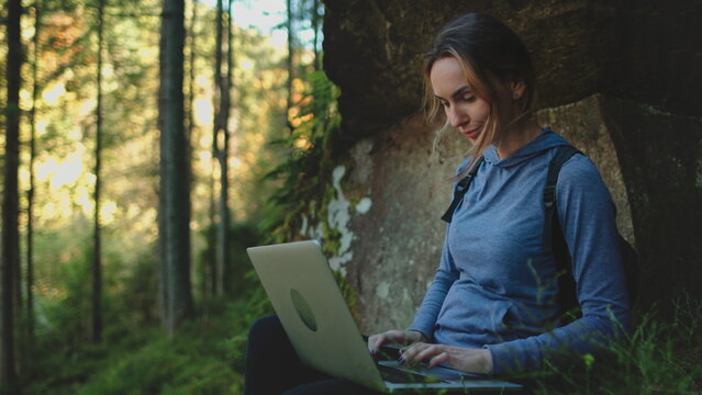 Woman enjoy working in wild nature landscape. Freelance girl typing on laptop while resting sitting under the tree in beautiful forest. Digital nomad remote work IT specialist, people travel, tourism
