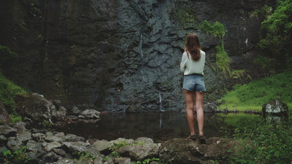 Woman enjoy jungle waterfall standing on rock. Travel to Bali island, Indonesia. Outdoor lifestyle travel on summer holiday vacation. Dark dramatic toning. Back view