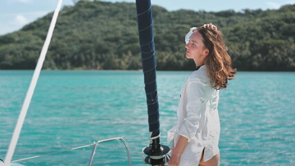 Woman in white shirt relaxes standing on yacht deck. Caucasian girl enjoys beautiful sea green...