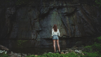 Drone shot panorama woman enjoy green jungle rock waterfall standing on stone back view. Girl relax outdoor lifestyle travel on summer holiday vacation. Dramatic dark toning.